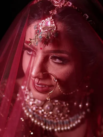 A close-up bridal portrait focusing on the bride's expressive eyes as she looks out from under her veil. This shot highlights the details of her makeup, the large nose ring (nath), and the ornate maang tikka, creating a captivating and traditional look.
