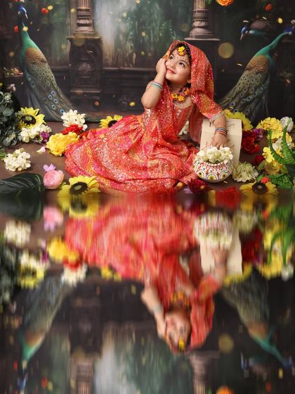 A beautiful portrait of a little girl dressed as Radha, lost in a divine dream. The reflection effect adds a layer of artistry to this image, set against a backdrop of peacocks and flowers.