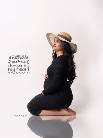 A stylish and modern solo portrait. The mom-to-be wears a simple black dress and a chic hat, looking up thoughtfully. This image has a contemporary and fashionable feel.