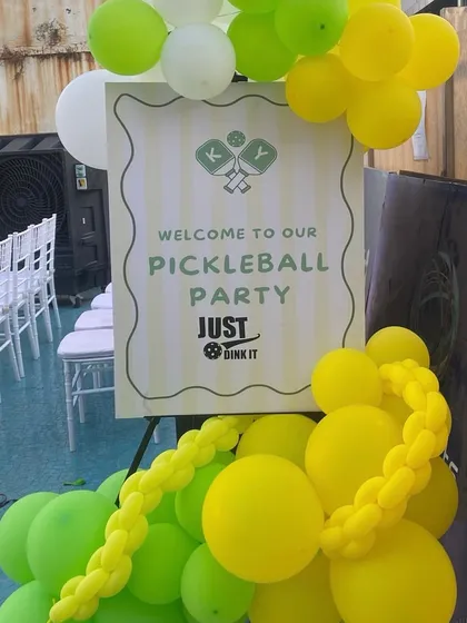 The welcome sign for the pickleball party, inviting guests to "Just Dink It." The sign is paired with a cheerful yellow and green balloon arrangement to set the tone for a day of fun.