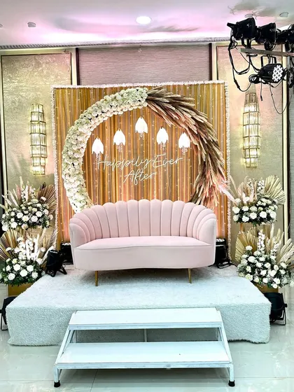 This is a seating area designed for a bohemian-themed Sangeet. The stage features a backdrop of gold fabric and dried palm leaves, with a "Happily Ever After" sign. The pink sofa and floral arrangements with pampas grass complete the chic, modern-boho look.