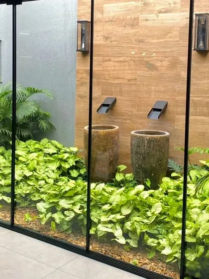 Bringing the calmness of water indoors. This modern courtyard features twin minimalist wall spouts cascading into rustic urns, surrounded by lush greenery. A floor-to-ceiling glass wall seamlessly integrates this piece of peace into the home's interior living space.