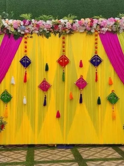 A bright and colorful backdrop perfect for a Haldi or Mehndi ceremony. This setup features a yellow curtain with pink and green drapes, decorated with traditional kite hangings.