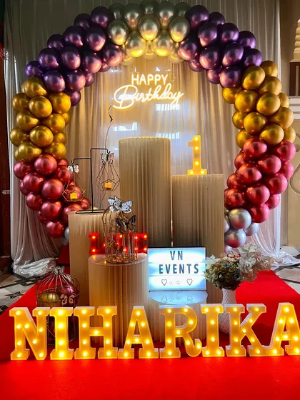 A first birthday decoration for 'NIHARIKA'. The setup features a rainbow-colored chrome balloon arch, ribbed pedestals, and bright marquee letters for the name.