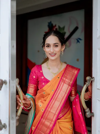 A welcoming and joyful portrait of the bride in a vibrant orange and pink saree. Her smile as she opens the doors creates a warm and inviting image, perfect for capturing the happiness of the occasion.