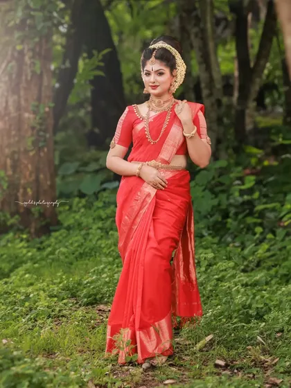 A full-length portrait set in a lush green forest. The vibrant red of the saree stands out beautifully against the natural backdrop, creating a striking contrast.