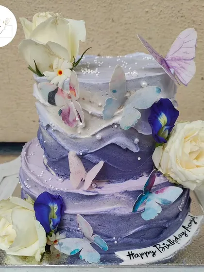 A stunning two-tier butterfly cake with a beautiful blueberry-purple ombre effect. The design is enhanced with fresh white roses, edible butterflies, and delicate blue pea flowers.