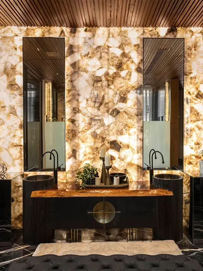 Here, a backlit onyx panel serves as a radiant feature wall behind the twin vanities. The warm, ethereal glow of the stone, paired with dark marble and wood ceilings, transforms the bathroom into a space of dramatic elegance.