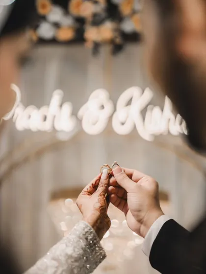 A creative close-up of the couple exchanging rings, framed against the blurred background of their names in neon lights. A symbolic and artistic shot.
