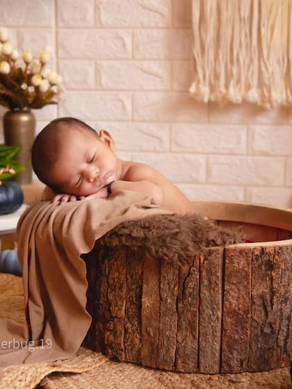 A close-up shot of a newborn sleeping peacefully in a rustic, bark-lined bowl. The warm, natural textures create a cozy and organic feel.