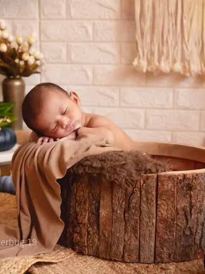 A close-up shot of a newborn sleeping peacefully in a rustic, bark-lined bowl. The warm, natural textures create a cozy and organic feel.