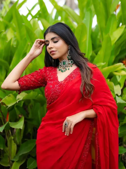 A striking portrait in a vibrant red saree. The lush green foliage in the background creates a beautiful contrast, making the color of the saree and the emerald jewelry stand out.