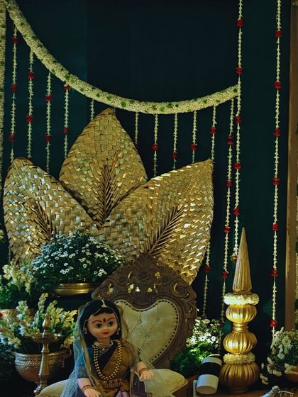 An elegant decor piece featuring a traditional throne against a dark green backdrop, accented with golden palm leaves and strings of jasmine flowers.
