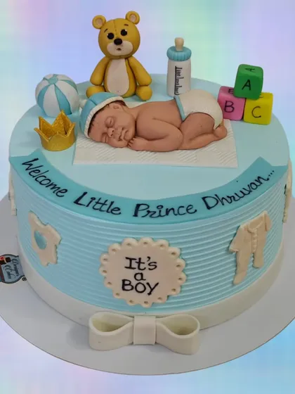 A "Welcome Little Prince" cake for a newborn baby boy. This design features a sleeping baby figurine, a teddy bear, and other cute baby items.