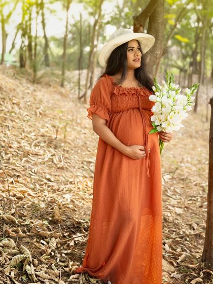 A serene moment in the forest. The mother-to-be, in a lovely terracotta-colored dress, holds a bouquet of white flowers, looking peaceful and connected to nature.