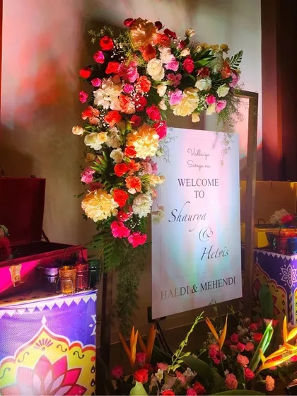 A personalized welcome station for a Haldi and Mehendi event, featuring a custom sign, colorful floral arrangements, and hand-painted decorative boxes.
