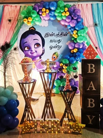 This is a custom Krishna-themed birthday party decoration. The setup features a playful illustration of little Krishna, peacock feather details, and a colorful balloon arch, making it a divine celebration for a child's special day.