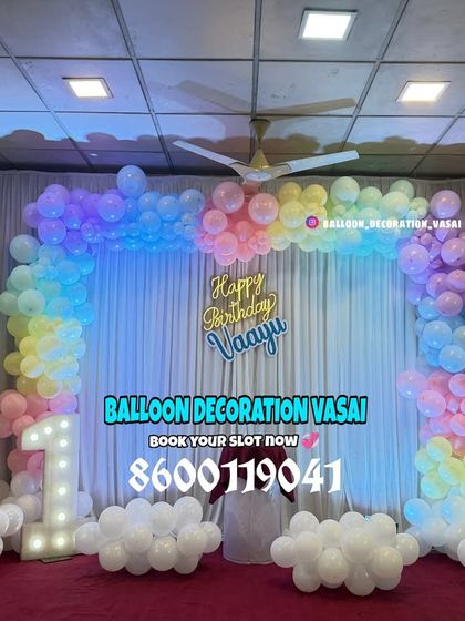 A dreamy pastel rainbow theme for Vanyu's first birthday. The balloon arch in soft colors against a white curtain with fairy lights creates a magical effect, complete with a marquee number '1'.