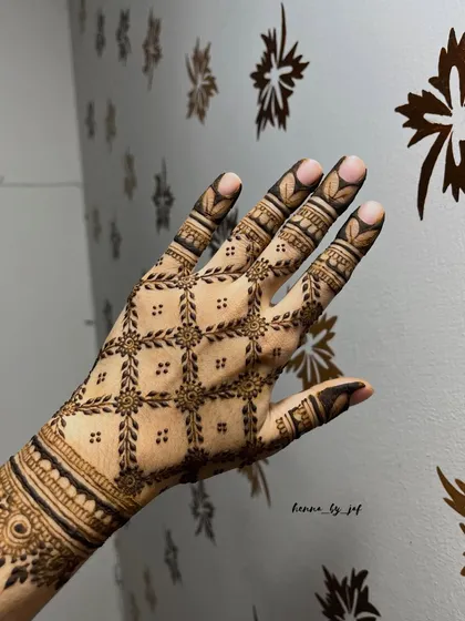A modern geometric henna design with a precise grid pattern covering the back of the hand. Each square contains a small floral motif, demonstrating skill in creating clean lines and symmetrical art.