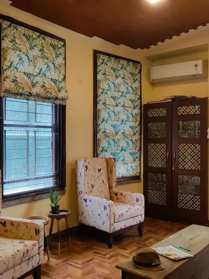 A quiet corner of a home where everything is in its place. The custom floral-patterned Roman blinds and matching wingback chairs create a cohesive and restful interior, complemented by the warm tones of the parquet flooring.