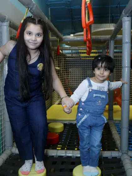 Teamwork makes the dream work! An older child helps a younger one navigate a netted bridge, showcasing the social interaction our play zones encourage.