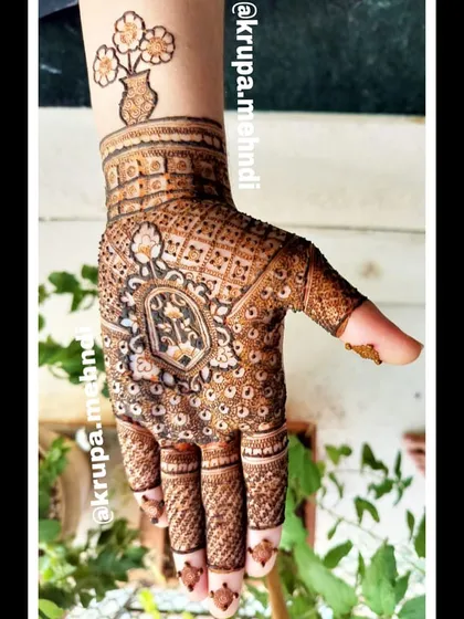A full, intricate design with a modern twist. The central motif is traditional, but the small tattoo-style flower pot on the wrist adds a unique touch.