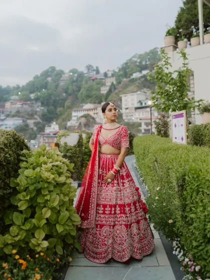 A full length portrait of the bride in her stunning red lehenga, set against the lush greenery and hillside architecture of Mussoorie. Her confident pose and elegant attire are the focus.
