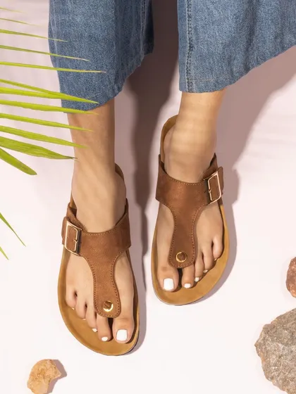 An on-model shot of brown T-strap sandals, styled with natural props like rocks and a palm leaf. This creates a casual, earthy, and comfortable look.