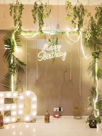 A beautiful bohemian-themed 60th birthday decoration. This elegant setup features a frame draped with greenery and fairy lights, a neon sign, and large marquee numbers.