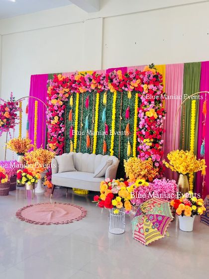 A lavish and colorful Mehendi setup in a large hall. The backdrop features a green panel with floral borders, surrounded by vibrant drapes, multiple floral arrangements, and traditional Rajasthani umbrellas.