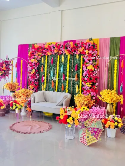 A lavish and colorful Mehendi setup in a large hall. The backdrop features a green panel with floral borders, surrounded by vibrant drapes, multiple floral arrangements, and traditional Rajasthani umbrellas.