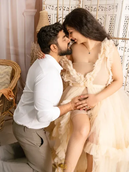 An intimate and playful moment between a couple. The partner kneels to embrace his wife, who is seated and laughing, both dressed in soft, neutral tones.