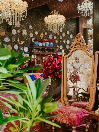 A gilded antique mirror and vanity stool are framed by lush plants and crystal chandeliers, creating a quiet, elegant corner within the bustling, vibrant event space.