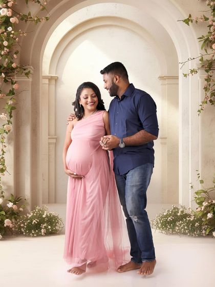 A candid moment of joy and connection. This couple shares a smile in a beautiful, light-filled studio setting with floral arches, creating a soft and romantic pregnancy portrait.