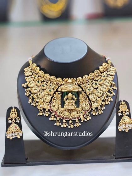 This stunning choker features a central pendant of Goddess Lakshmi in her temple, surrounded by intricate peacock carvings. It comes with matching jhumka earrings.