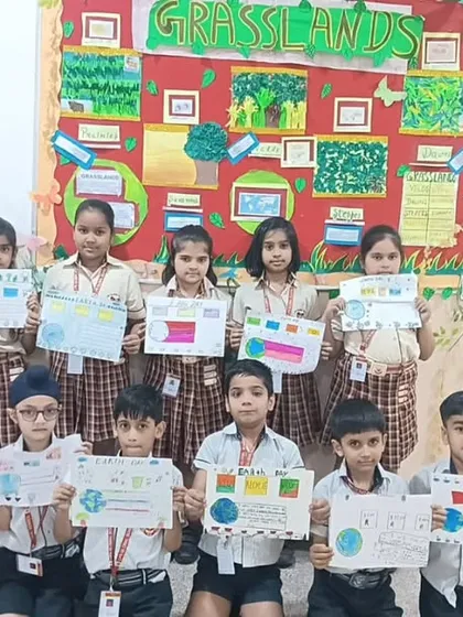 A group of students poses with their Earth Day posters in front of a bulletin board themed on grasslands, showcasing their classroom learning on ecosystems.