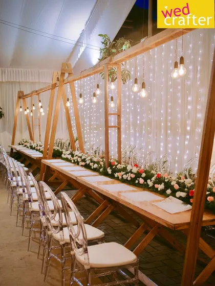 Elegant long wooden tables paired with modern transparent ghost chairs, set under hanging Edison bulbs against a backdrop of fairy lights.