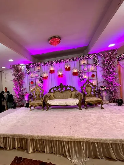An elegant reception stage with a white backdrop featuring decorative birdcages and floral arrangements. The purple uplighting adds a touch of modern glamour to the classic and regal seating.