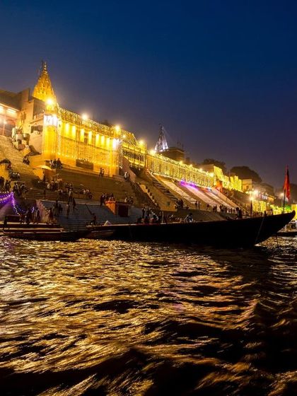 The ghats of Varanasi illuminated for Dev Deepawali, as seen from the river. The golden light reflecting on the water creates a stunning and serene cityscape.