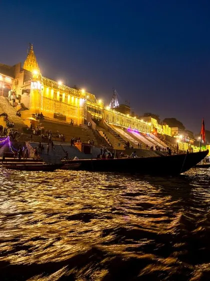 The ghats of Varanasi illuminated for Dev Deepawali, as seen from the river. The golden light reflecting on the water creates a stunning and serene cityscape.