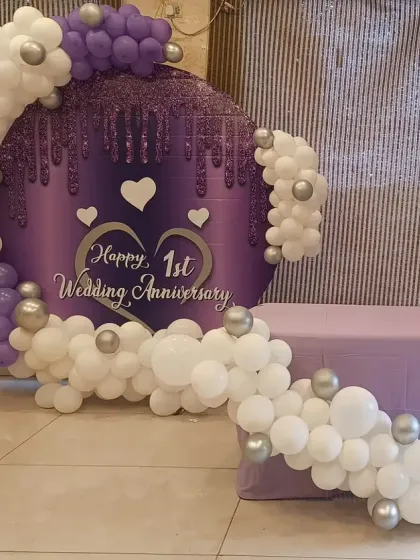 An elegant first wedding anniversary setup with a custom purple backdrop and a white and silver balloon garland.