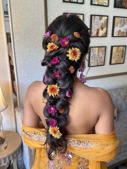 A close-up of a vibrant Haldi braid, filled with bright yellow and pink fresh flowers. The texture is messy and romantic, perfect for the fun and festive atmosphere of a Haldi ceremony.