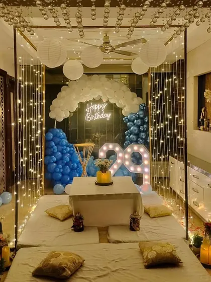 An intimate and cozy indoor birthday dinner setup. This features a cabana frame draped with fairy lights, low seating with plush cushions, and a balloon backdrop in shades of blue and white, perfect for a surprise celebration at home.
