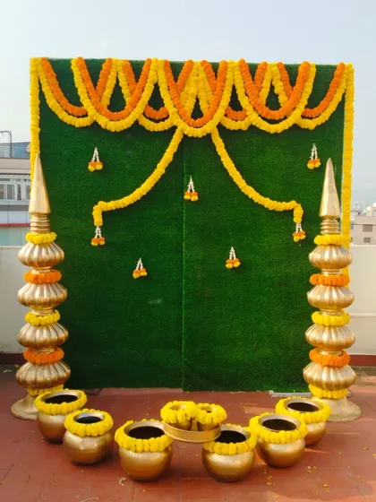 A simple and elegant Haldi backdrop with a green base and marigold garlands. The setup includes brass pots for a traditional touch.