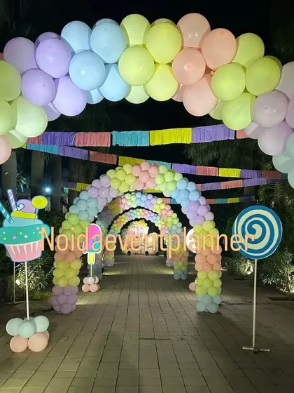 An immersive candyland party entrance featuring a long tunnel of pastel rainbow balloon arches and cute lollipop and cupcake props along the walkway.