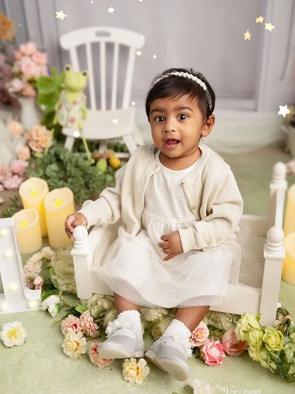 A classic first birthday portrait. The little girl sits on a miniature bench surrounded by soft flowers and candles, creating a timeless and elegant memory.