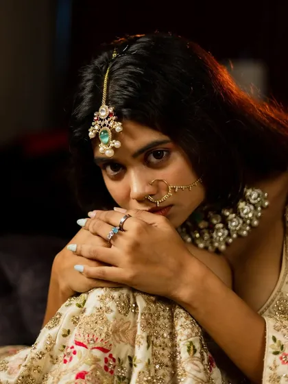 An intimate and artistic bridal portrait. The bride's pose, with her hands framing her face, draws attention to her expressive eyes and traditional maang tikka and nath.
