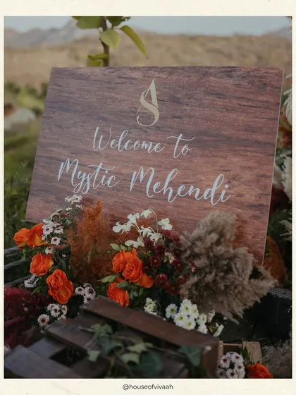 A rustic wooden welcome sign for a "Mystic Mehendi," framed with pampas grass and burnt orange florals, setting an earthy and bohemian tone for the event.