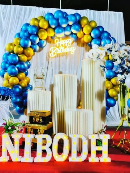 A striking blue and gold theme set against a white drape backdrop. This setup includes a balloon arch, neon sign, and light-up marquee letters spelling 'NIBODH' on a red carpet.