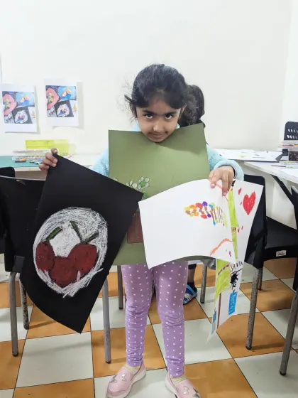 Uvika from the elementary batch proudly displays her work. Her projects include a study of cherries on a dark background and other imaginative drawings.
